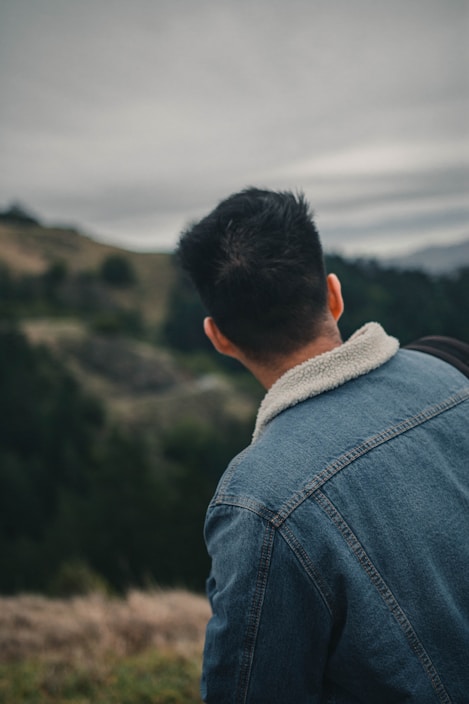 A person wearing a denim jacket with a fluffy collar stands outdoors, looking towards a scenic landscape with rolling hills and a cloudy sky. The scene appears calm and natural.