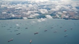 Aerial view of numerous cargo ships scattered across a large body of water near a coastline. Puffy white clouds hover over the scene, casting soft shadows on the water. In the distance, urban landscape with clusters of buildings and green areas is visible.