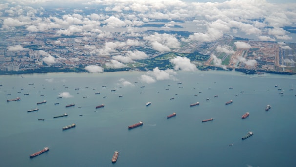 Aerial view of numerous cargo ships scattered across a large body of water near a coastline. Puffy white clouds hover over the scene, casting soft shadows on the water. In the distance, urban landscape with clusters of buildings and green areas is visible.