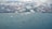 Aerial view of numerous cargo ships scattered across a large body of water near a coastline. Puffy white clouds hover over the scene, casting soft shadows on the water. In the distance, urban landscape with clusters of buildings and green areas is visible.
