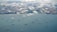 Aerial view of numerous cargo ships scattered across a large body of water near a coastline. Puffy white clouds hover over the scene, casting soft shadows on the water. In the distance, urban landscape with clusters of buildings and green areas is visible.