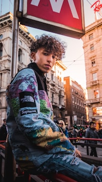 A person with curly hair is wearing a colorful jacket featuring world map patterns, leaning against a railing in an urban environment. The street is bustling with people walking, and there are historic buildings in the background. A large red and white sign overhead suggests a metropolitan setting during daytime.