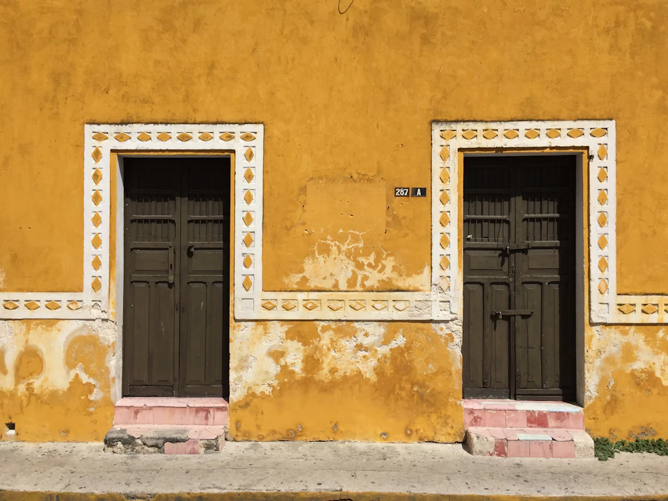 The image shows a two-story building with yellow walls and black doors, both adorned with white designs on the lower half. The upper floor features a smaller door with a window, while the lower floor has a larger door without any visible windows. The building is situated on a street corner, with a sidewalk running parallel to it in front of the structure.