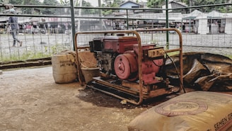 A portable generator surrounded by construction materials, including bags and a large container. The setting appears to be an open-air market or construction site with people in the background and various stalls or small buildings visible beyond a metal fence.