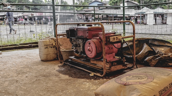 A portable generator surrounded by construction materials, including bags and a large container. The setting appears to be an open-air market or construction site with people in the background and various stalls or small buildings visible beyond a metal fence.
