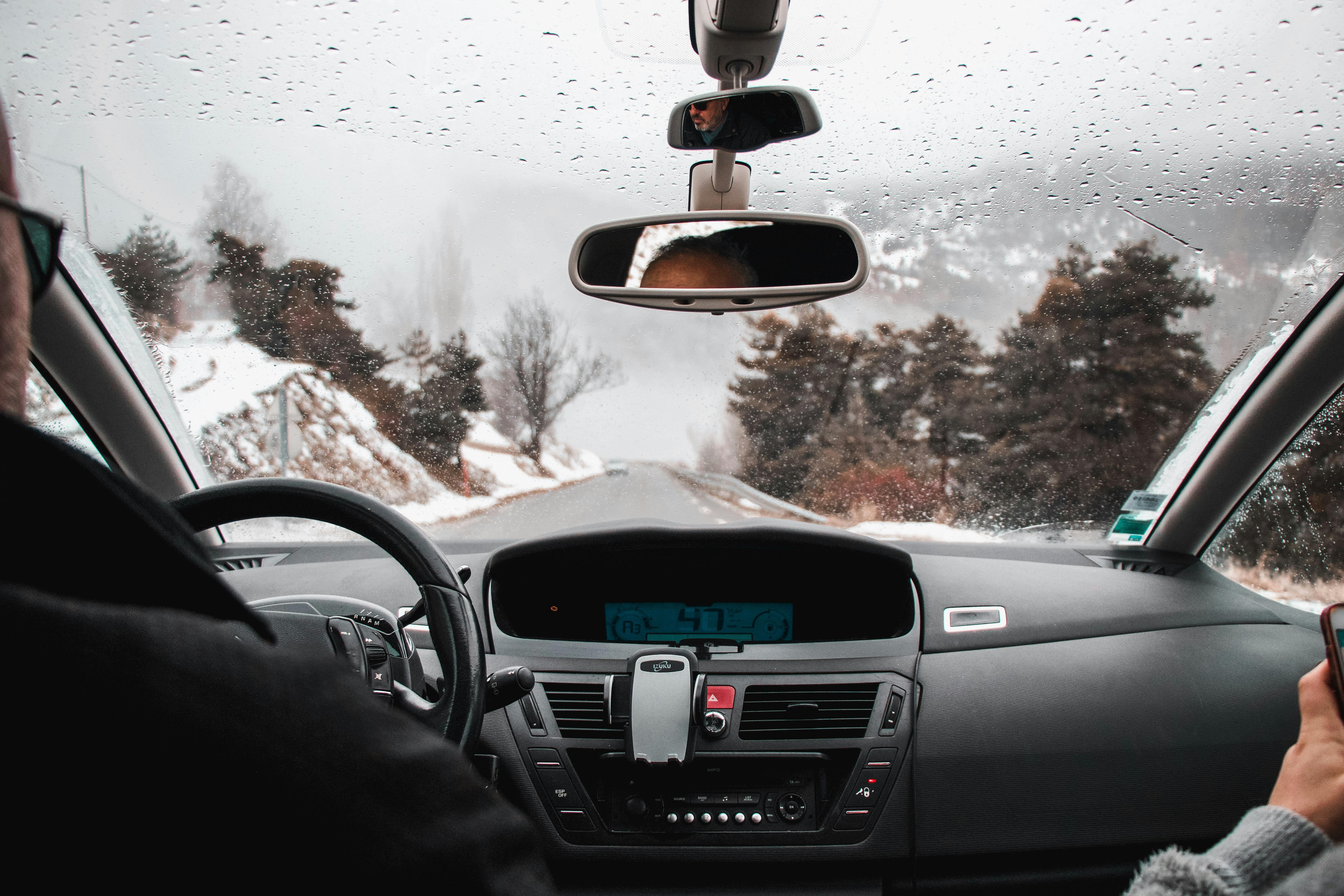 Candid photograph from the back seat capturing the dashboard and windshield with a snow-covered road ahead. The frame emphasizes the quiet winter scene framed by the car's interior.