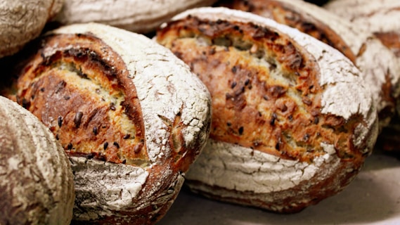 Several loaves of artisanal bread with a rustic, golden-brown crust. The bread features a dusting of flour and visible seeds embedded in the crust, creating a textured appearance.