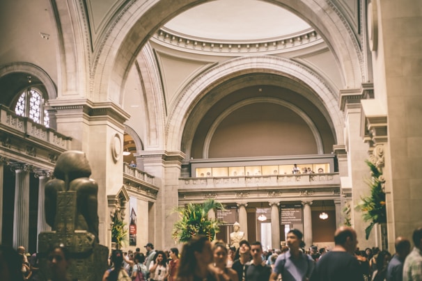 A large, grand hall with ornate architectural details, including arched ceilings and classical columns. The space is filled with people, indicating a busy or popular setting. The hall features a statue in the foreground and additional sculptures or exhibits are visible in the background.