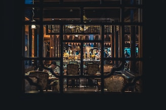 An intimate setting inside a dimly lit restaurant with ornate decorations. Tables and chairs are arranged around a central area, where several people are seen conversing or dining. Wooden structures and decorative elements lend an elegant and traditional atmosphere.