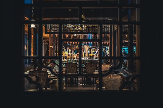 An intimate setting inside a dimly lit restaurant with ornate decorations. Tables and chairs are arranged around a central area, where several people are seen conversing or dining. Wooden structures and decorative elements lend an elegant and traditional atmosphere.