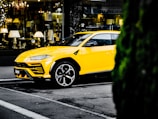 A bright yellow luxury SUV is parked on the street in an urban setting. The vehicle is positioned in front of a store window displaying various elegant lighting fixtures and home decor items. The surrounding environment includes typical city street elements and a glimpse of greenery on the right side.