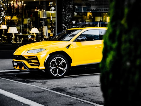 A bright yellow luxury SUV is parked on the street in an urban setting. The vehicle is positioned in front of a store window displaying various elegant lighting fixtures and home decor items. The surrounding environment includes typical city street elements and a glimpse of greenery on the right side.