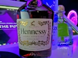 A close-up of a bottle of Hennessy cognac is prominently displayed, flanked by a bottle of vodka and Patron tequila. The background features a digital screen with purple neon lights and text partially visible, creating a club or bar-like atmosphere.