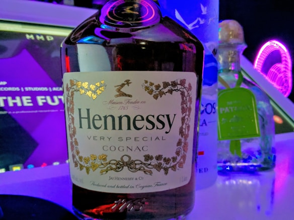 A close-up of a bottle of Hennessy cognac is prominently displayed, flanked by a bottle of vodka and Patron tequila. The background features a digital screen with purple neon lights and text partially visible, creating a club or bar-like atmosphere.
