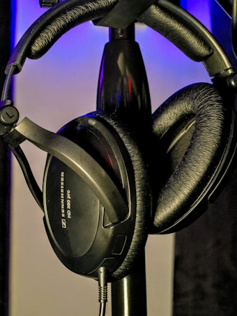 A pair of black over-ear headphones is hanging on a stand. The background features muted lighting with a bluish tint, creating a subtle contrast with the dark headphones. The headphones have plush ear cushions and an adjustable headband.