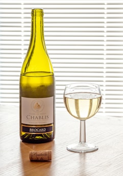A bottle of Chablis wine labeled Pierre de Préhy, accompanied by a glass filled with white wine and a cork. They are placed on a wooden surface with blinds in the background, allowing soft light to create an inviting atmosphere.