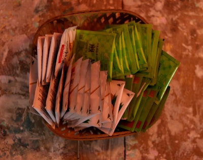 A basket filled with packets of tea or drink mix, arranged with alternating green and white packaging. The basket is set on a textured, possibly wooden surface, with a warm lighting casting an orange hue over the scene.