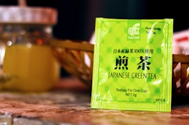 A brightly colored packet of Japanese green tea is prominently displayed in the foreground. The packet features green polka dots and Japanese characters, along with English text indicating it's a single-serving teabag. In the background, there is a glass jar with a metallic lid, slightly out of focus.