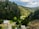 A scenic valley features lush green forests on rolling hills. A winding road cuts through the landscape, leading to a quaint house with a light blue roof nestled among trees. The scene is bathed in natural light, with clouds partially covering a blue sky.