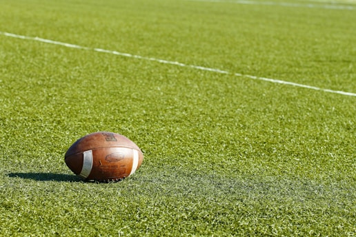 A football rests on a green field marked with white lines. The ball is positioned on the grass, with shadows suggesting sunlight.