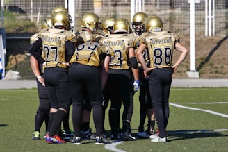 A group of American football players wearing matching gold and black uniforms and helmets, huddled together on a grassy field. The players have numbers on their jerseys and stand closely together, likely discussing a strategy. The background includes a fenced area and some natural surroundings.