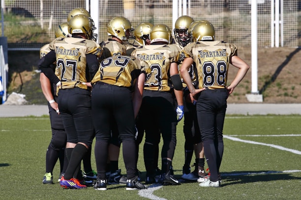 A group of American football players wearing matching gold and black uniforms and helmets, huddled together on a grassy field. The players have numbers on their jerseys and stand closely together, likely discussing a strategy. The background includes a fenced area and some natural surroundings.
