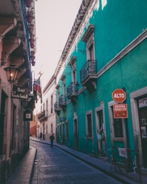 A narrow, cobblestone street flanked by colorful buildings, one of which is painted green with ornate balconies. There is a stop sign labeled 'ALTO' in Spanish, indicating a pedestrian zone with restricted access. A few people can be seen walking along the street, creating a sense of everyday life in a historical urban setting.