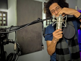 A person with curly hair and glasses is adjusting a microphone in a recording studio. The individual is wearing a blue shirt and a black-and-gray checkered scarf. The studio is equipped with soundproof padding and several microphone stands.