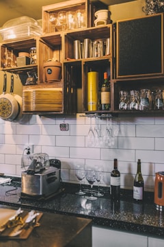 A cozy kitchen counter with a black speckled countertop, decorated with a series of wooden shelves displaying various kitchen items including glassware, bottles of wine, metal containers, and a toaster. The white subway tile backsplash adds a clean and bright contrast to the warm tones of the wooden shelves. Hanging wine glasses are neatly arranged beneath the shelves, and there are a few more wine bottles placed on the counter.