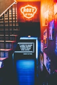 A dimly lit stairway leading down to a venue, illuminated by a bright orange neon sign reading 'Roxy LP Pub'. The wall to the right is adorned with various posters and flyers. Below the neon sign, a blackboard displays a message in white text expressing a focus on making women happy.