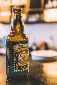 A glass bottle of 'Great White Beer' with a label featuring a cartoon shark holding a surfboard. The background is a softly focused, warm-toned kitchen or bar setting, with out-of-focus plates and soft lighting.