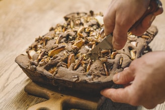 A freshly baked pie with a golden-brown crust is being sliced with a knife. The pie contains pieces of fruits and nuts visible on top. It is placed on a wooden board, and the person cutting it has both hands positioned firmly.