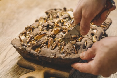 A freshly baked pie with a golden-brown crust is being sliced with a knife. The pie contains pieces of fruits and nuts visible on top. It is placed on a wooden board, and the person cutting it has both hands positioned firmly.