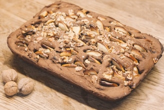 A rectangular baked dessert featuring a dense looking cake filled with chopped nuts and slices of fruit. The cake appears to be freshly baked and is placed on a wooden surface, with two whole walnuts nearby.