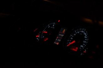 A dimly lit car dashboard displaying illuminated speedometer and tachometer gauges with red and white indicators. The central digital display shows a temperature of 21°C and a passenger safety alert icon.