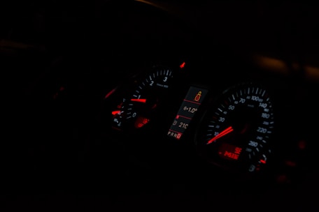 A dimly lit car dashboard displaying illuminated speedometer and tachometer gauges with red and white indicators. The central digital display shows a temperature of 21°C and a passenger safety alert icon.