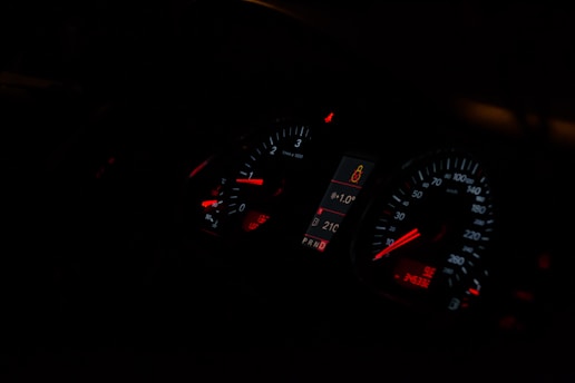 A dimly lit car dashboard displaying illuminated speedometer and tachometer gauges with red and white indicators. The central digital display shows a temperature of 21°C and a passenger safety alert icon.