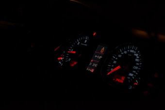 A dimly lit car dashboard displaying illuminated speedometer and tachometer gauges with red and white indicators. The central digital display shows a temperature of 21&deg;C and a passenger safety alert icon.