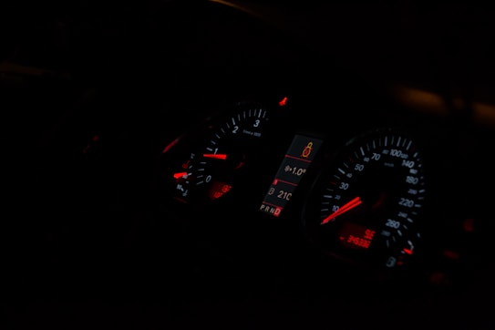 A dimly lit car dashboard displaying illuminated speedometer and tachometer gauges with red and white indicators. The central digital display shows a temperature of 21°C and a passenger safety alert icon.