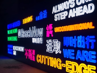 Neon signs featuring various motivational words and phrases in English and Chinese. The signs display messages like 'Reach Now,' 'Unconventional,' 'Cutting-Edge,' and 'Always One Step Ahead' in bright colors against a dark background.
