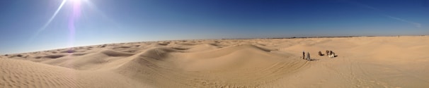 Expansive sand dunes stretch across the landscape under a clear blue sky, with several people and camels visible in the distance, creating tracks on the sand.