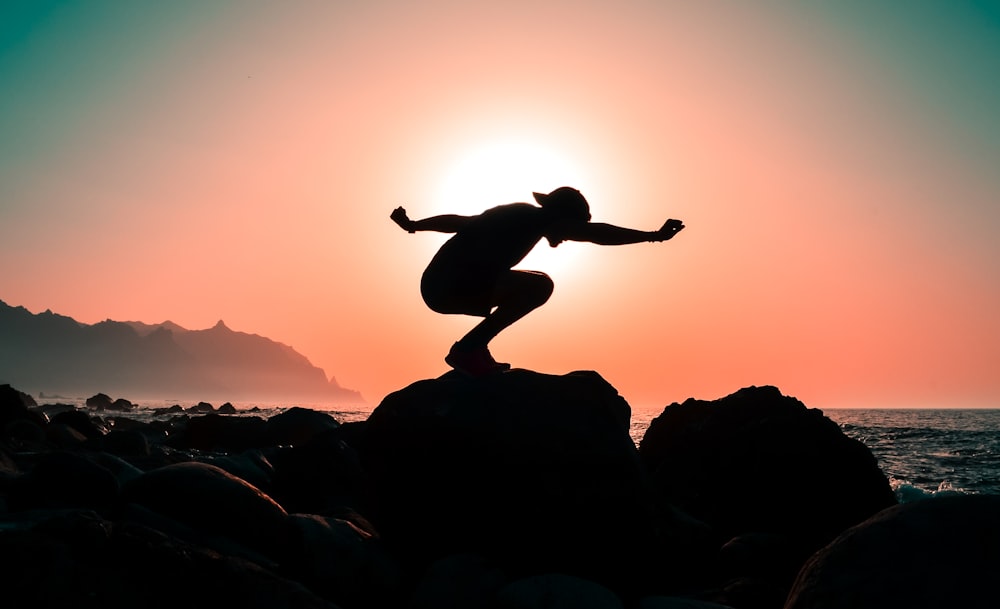 silhouette photography of person squatting on rock near body of water during golden hour