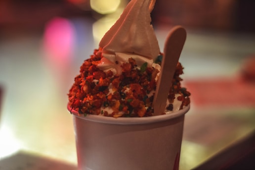 A cup of soft-serve ice cream topped with crushed nuts and a wooden spoon inserted into it. The background is blurred, creating a bokeh effect with warm colored lights.