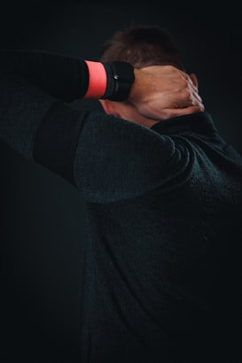 A person is seen from behind, wearing a dark sweater and an orange wristband, with hands placed on the back of their neck.