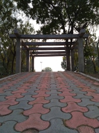 A pathway made of interlocking red and gray tiles leads to a pergola supported by concrete pillars. The path is surrounded by lush green trees and the sky is visible through the branches.