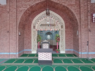 An ornate architectural interior features a detailed archway constructed with red bricks and embellished with intricate white carvings. The floor is covered with a patterned green carpet. Floral arrangements and decorations are placed on both sides of a central raised platform with a decorative chair.
