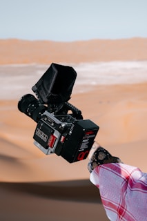 A person holds a professional video camera against a backdrop of sandy desert dunes. The individual's hand is partially visible, wearing a patterned sleeve. The camera features a large lens and a viewfinder covered with a protective hood.