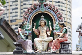 Three intricately carved and painted statues are depicted in a traditional Hindu style. The central figure is a seated deity adorned with jewelry and holding various traditional symbols, surrounded by an ornate arch. Flanking the deity are two attendant figures, also sitting and holding offerings or symbols. The background features a modern building, contrasting the traditional statues.
