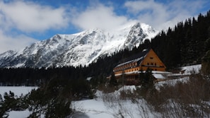 A large, wooden lodge sits nestled among snow-covered trees in a mountainous landscape. The tall, rugged mountains in the background are blanketed with snow, under a sky partially covered with clouds. The building has a steep, dark roof and numerous windows, suggesting a welcoming retreat in a serene, natural setting.