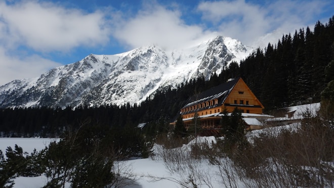 A large, wooden lodge sits nestled among snow-covered trees in a mountainous landscape. The tall, rugged mountains in the background are blanketed with snow, under a sky partially covered with clouds. The building has a steep, dark roof and numerous windows, suggesting a welcoming retreat in a serene, natural setting.